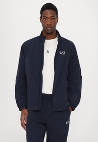 Giacca zip-up blu marina con polsini elastici, tessuto leggero e logo frontale. Indossata sopra una camicia bianca, abbinata a pantaloni coordinati.