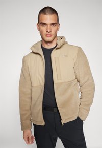 Beige Fleecejacke mit weicher Textur, verfügt über eine Kapuze, einen Reißverschluss vorne und eine Brusttasche. Schwarzes Untershirt und dunkle Hose runden den Look ab.