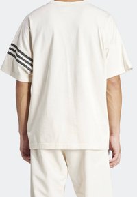 Cream-colored cotton oversized t-shirt with short sleeves and black striped accents on the left shoulder. No visible graphics or patterns.