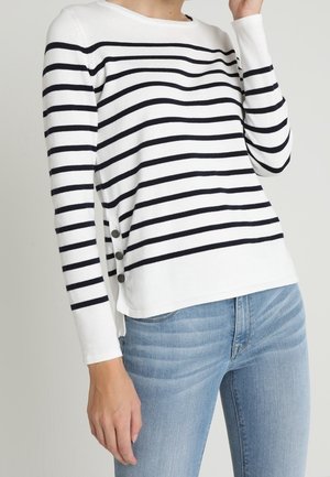 Mujer que lleva una camiseta blanca de manga larga con rayas horizontales negras y tres botones en el lateral, combinada con vaqueros azul claro.