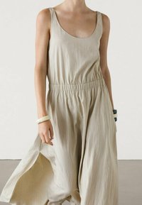 Robe longue beige sans manches avec un décolleté arrondi et une taille froncée. Tissu léger et texturé avec une silhouette fluide.