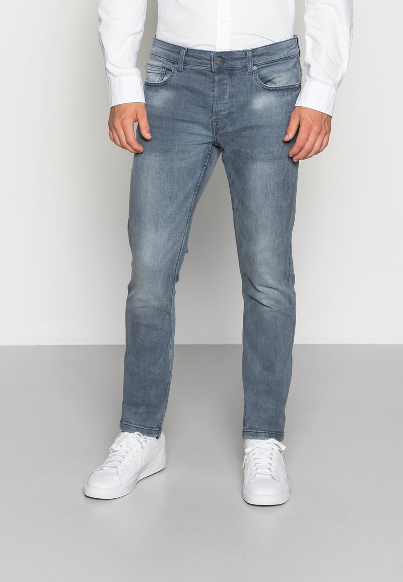 Homme portant un jean bleu délavé slim fit, une chemise blanche boutonnée et des baskets blanches, debout sur un sol gris devant un fond uni.