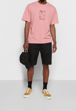 Man draagt een roze T-shirt met print, zwarte korte broek, zwarte sokken, mosterdgele sneakers, houdt een zwarte honkbalpet in één hand vast.