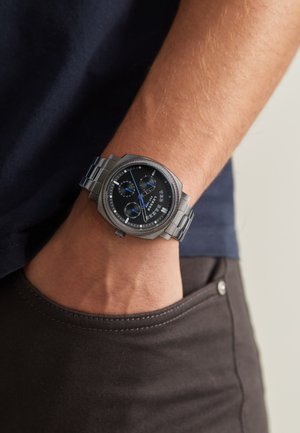 Reloj cronógrafo plateado con un dial negro texturizado, acentos azules, tres subdiales y una pulsera de eslabones metálicos. Llevarlo en una muñeca.