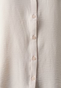 Camicia button-up beige chiaro realizzata in tessuto testurizzato. Presenta cinque bottoni tondi di colore chiaro lungo la parte frontale. Aspetto morbido e sgualcito.