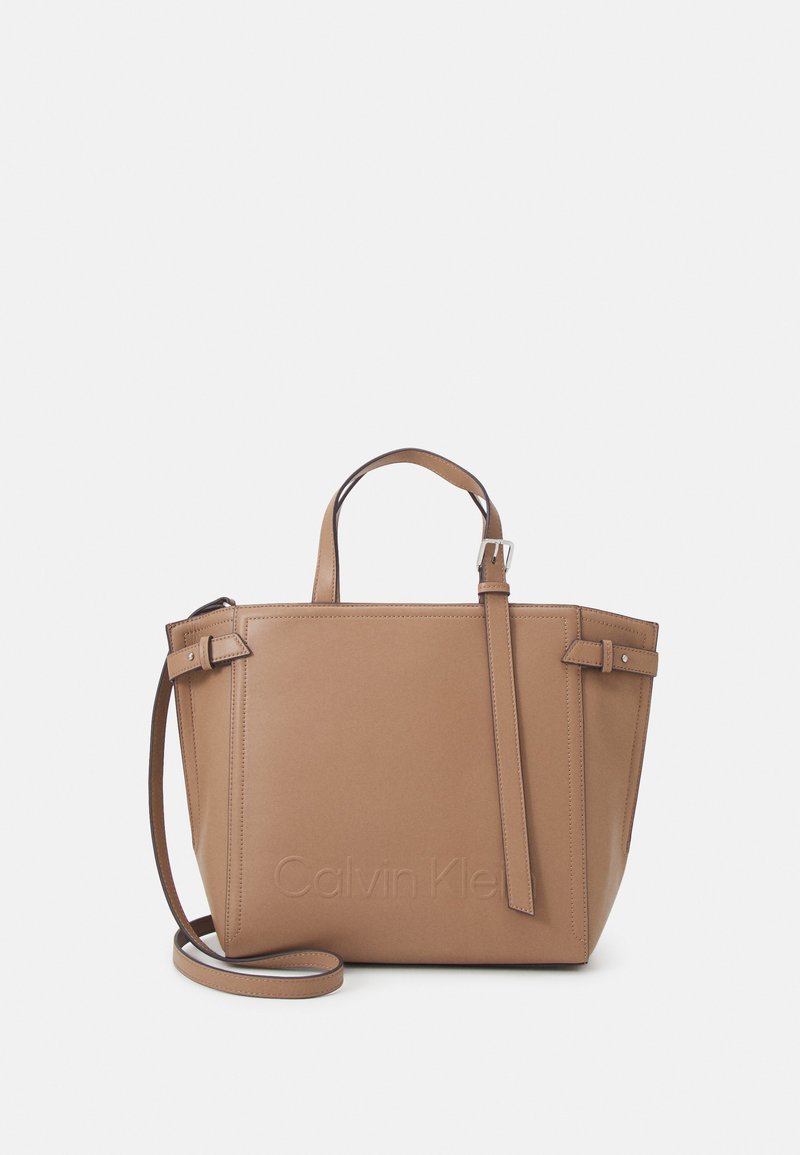 Sac à main en cuir tan avec deux poignées et une bandoulière réglable. Présente une forme rectangulaire, un marquage en relief et un design minimaliste.