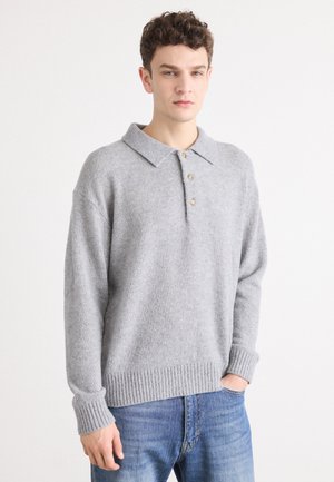 Hellgrauer Strickpullover mit Kragen, drei Knöpfen und gerippten Bündchen sowie Saum. Über blauen Jeans getragen.