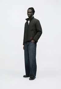 Black quilted jacket with high collar and zipper, featuring a left pocket and red logo. Paired with loose-fit blue denim jeans and black shoes.