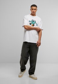T-shirt blanc à manches courtes avec un graphique coloré d'un personnage de dessin animé, associé à un pantalon ample gris foncé et des baskets beiges.