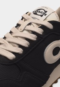 Κοντινό πλάνο μαύρου και λευκού αθλητικού παπουτσιού Coach με εκρού κορδόνια, υφασμάτινο πάνω μέρος και λογότυπο στη γλώσσα και την πλαϊνή πάνελ.