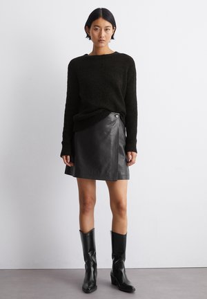 Schwarzer Strickpullover mit langen Ärmeln, kombiniert mit einem eng anliegenden schwarzen Lederrock, der über einen seitlichen Knopfdetail verfügt. Schwarze kniehohe Stiefel runden den Look ab.