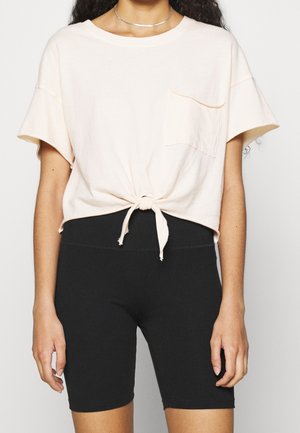Vrouw draagt een lichtroze shirt met korte mouwen en een strik aan de voorkant, samen met zwarte aansluitende fietsbroeken tegen een effen achtergrond.