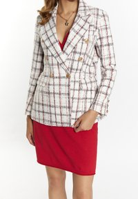 Blazer en tweed blanc avec un motif écossais rouge et noir, design double boutonnage, boutons dorés et coupe structurée. Associé à une robe rouge.