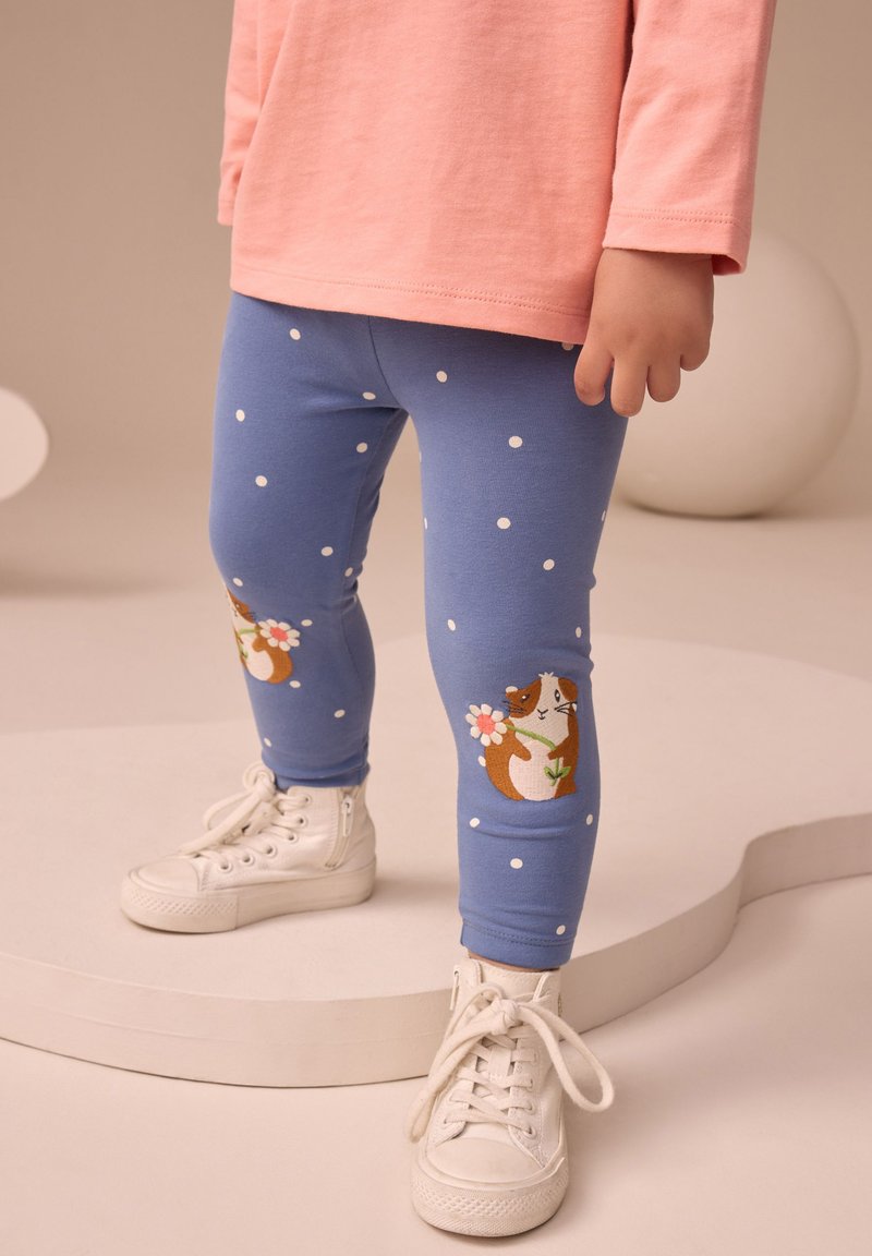 Leggings en coton bleu à pois blancs avec un motif de cochon d'Inde tenant une fleur sur le genou gauche, assortis à des baskets blanches montantes.