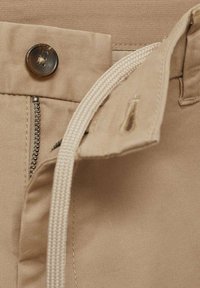 Béžová látka s viditelným kovovým zipem, zapínáním na knoflík a bílým šňůrkovým stažením. Textura je hladká s čistými detaily šití.