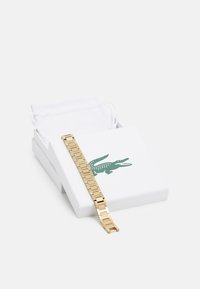 Bracelet en métal doré avec des maillons carrés, présenté dans une boîte blanche arborant un logo de crocodile vert et accompagné d'un sac blanc.