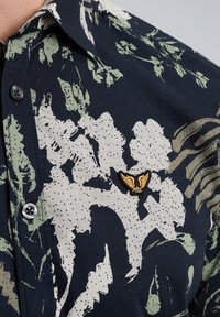 Camisa de manga corta en azul marino oscuro con un estampado floral en verde claro y crema, que presenta un pequeño logo bordado en dorado en el pecho.
