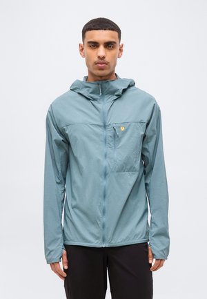 Νέος άνδρας που φοράει ανοιχτό μπλε μπουφάν με κουκούλα, φερμουάρ και τσέπη στο στήθος, στέκεται μπροστά σε απλό ανοιχτό φόντο.