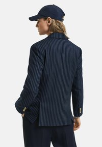 Blazer navy a righe con spalle strutturate, rever eleganti e dettagli in bottone dorato sui polsini. Completa un outfit coordinato.
