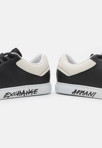 Svarte og hvite sneakers med overdel i teksturert lær, hvit gummisåle og grafiske tekstlogoer "EXCHANG" og "ARMANI" på siden.