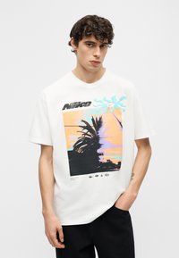 Νέος άνδρας με σγουρά μαλλιά που φοράει λευκό t-shirt Nike με γραφικό απεικόνιση ηλιοβασιλέματος και φοίνικα, στέκεται με το ένα χέρι στην τσέπη.