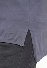 Szara koszulka z krótkim rękawem wykonana z miękkiej tkaniny, z rozcięciem po bokach u dołu i luźnym krojem. W zestawie z ciemnymi spodniami dżinsowymi.