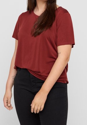 Femme portant un haut manches courtes rouge rouille et un pantalon noir, avec des tatouages visibles sur les bras, debout devant un fond clair et uni.