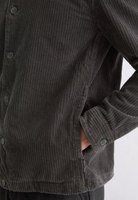 Dark grey corduroy shirt with vertical ribbed texture, button front closure, and side pocket, featuring a stitched label along the hem.