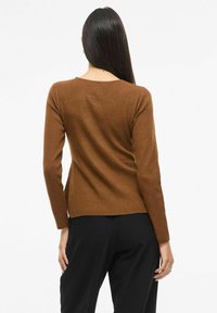 Pruun sooniline pikakäisline pluus ümarate kaelusega, keha jälgiv disain ja kerge kitsenemine allosas. Kandetud mustade pükstega.