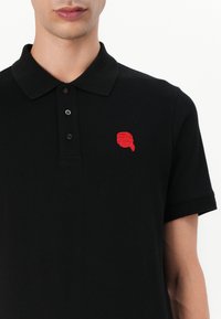 Μαύρο πόλο μπλουζάκι από βαμβάκι, με τρία κουμπιά στο κολάρο. Διακριτό κόκκινο κεντημένο λογότυπο στην αριστερή πλευρά του στήθους. Κοντά μανίκια.