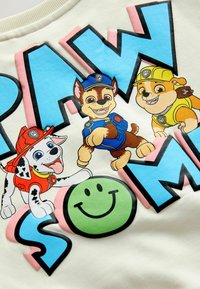 Trois chiots animés en uniformes de pompier, de policier et de construction avec de grandes lettres bleues et un visage souriant vert sur un tissu blanc.