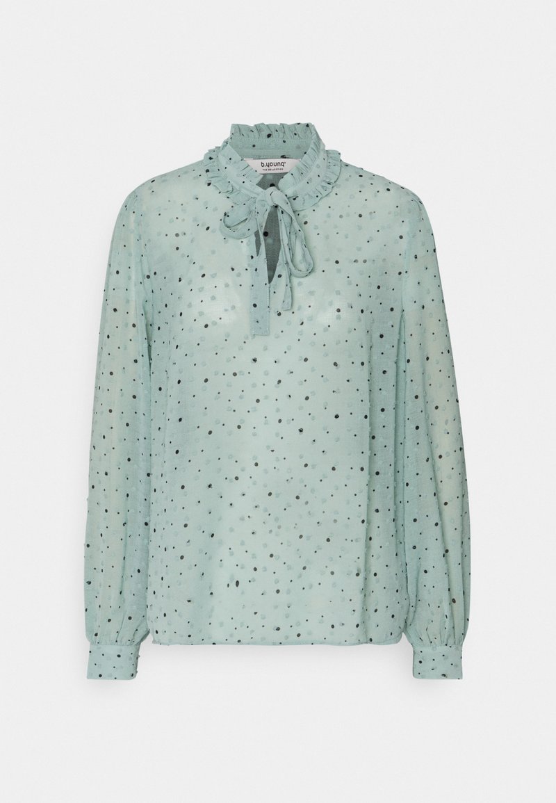 Blusa di un verde teal chiaro con scollatura a volant e dettaglio con fiocco. Realizzata in tessuto trasparente con pois neri sparsi, presenta maniche lunghe con polsini.