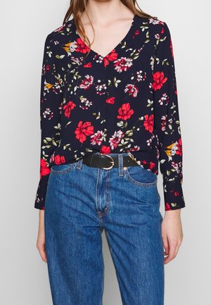 Kvinde iført mørk blomsterlangærmet bluse stukket ned i blå jeans med sort bælte, stående mod en ensfarvet lys baggrund.