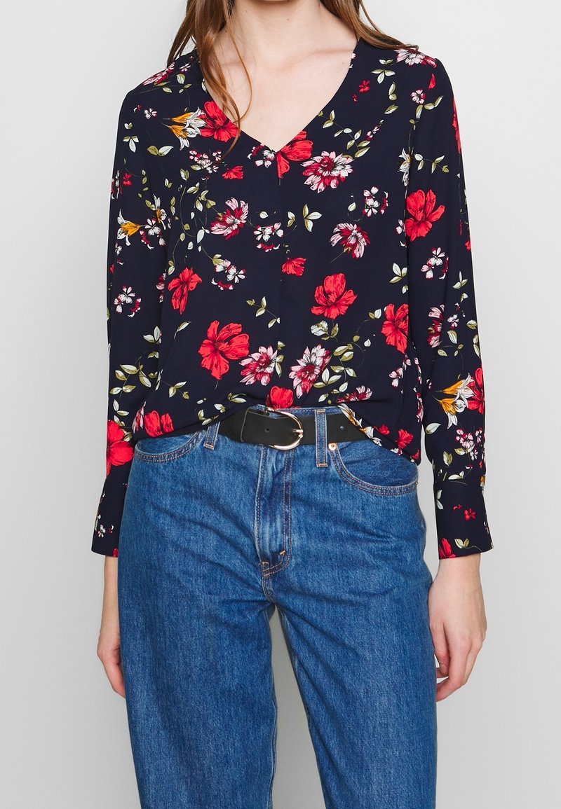 Femme portant un chemisier à manches longues à motif floral foncé, rentré dans un jean bleu avec une ceinture noire, debout devant un fond clair uni.