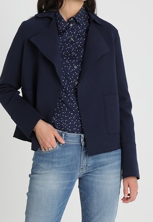 Kvinde iført en åben jakke i marineblå over en mønstret navyblå skjorte med knapper og lyseblå denimjeans.