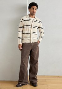 Bēša adīta kardigāna ar tumši ziliem svītrām, pogu aizdare, ar apkakli; kombinēts ar brūnām plašām biksēm un tumšiem apaviem. Koka grīdas fons.