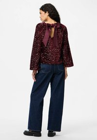 Top bordeaux con paillettes, maniche lunghe e chiusura a fiocco sul retro, abbinato a jeans in denim scuro a vita alta e gamba larga, e scarpe nere.