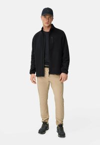 Schwarze Fleece-Jacke mit vollem Reißverschluss, getragen über einem schwarzen Hemd, kombiniert mit khakifarbenen Hosen und schwarzen Wanderschuhen. Mütze mit Logo.