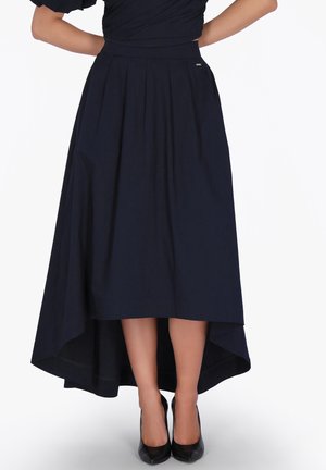 Femme portant une jupe midi bleu marine asymétrique plissée, associée à des chaussures à talons noirs, debout les mains sur les hanches.