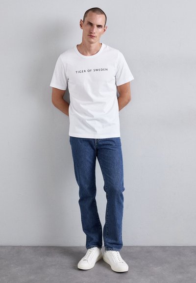Mann med kort hår står mot en ensfarget vegg iført en hvit "Tiger of Sweden" t-skjorte, blå jeans og hvite joggesko.