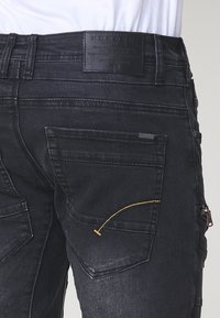 Pantalones de denim negro con un acabado texturizado, adornados con costuras amarillas, una etiqueta de marca de cuero y múltiples bolsillos.
