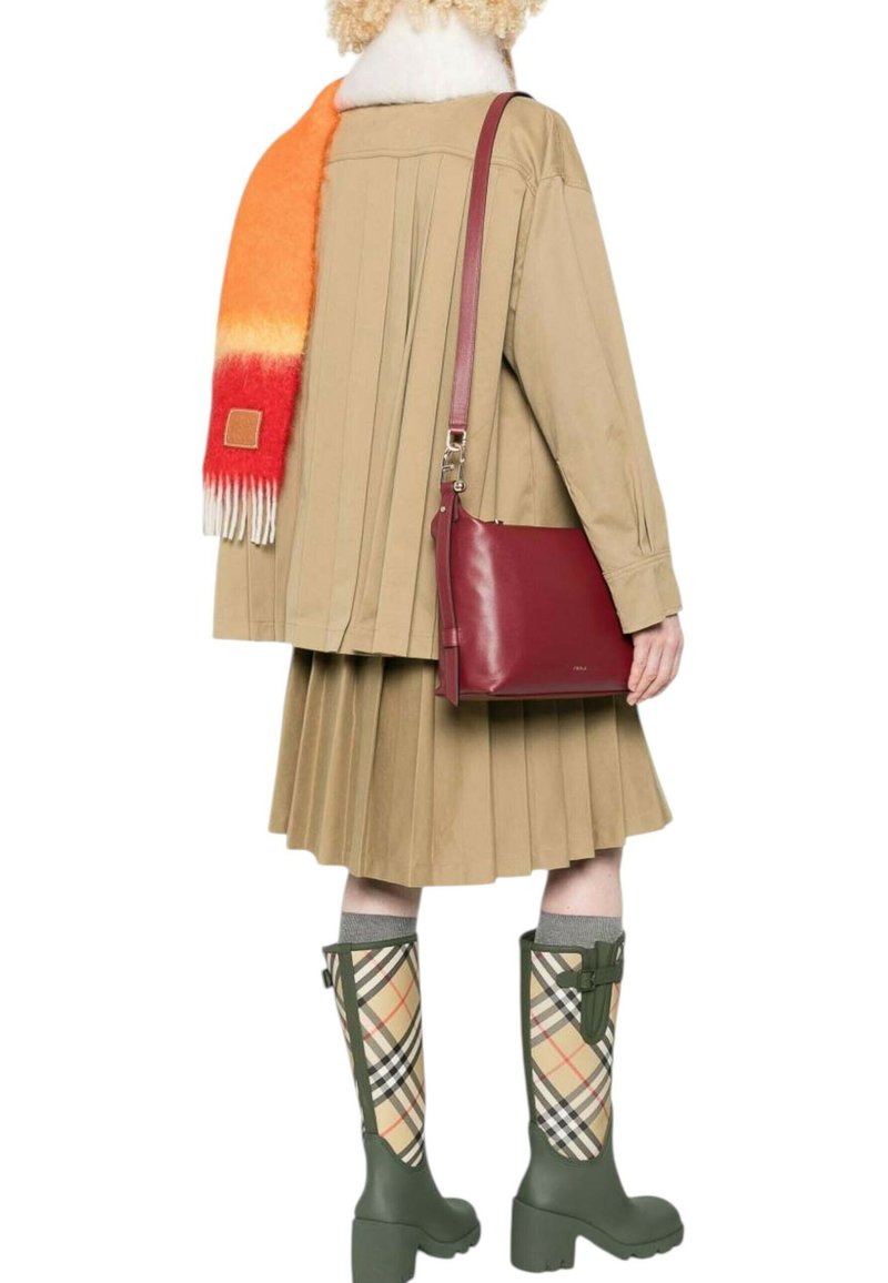 Persona che indossa un cappotto plissettato beige e una gonna, una sciarpa arancione-rossa, porta una borsa a tracolla bordeaux e indossa stivali al ginocchio a quadri verdi.