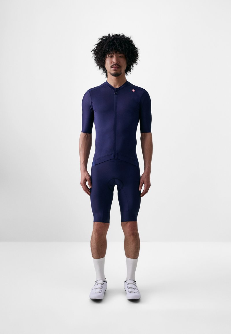 Marinefarbenes Radfahrerset aus dehnbarem Stoff, mit kurzen Ärmeln, Reißverschluss und gepolsterten Shorts. Weiße Socken und Radschuhe vervollständigen das Outfit.