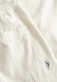 Gros plan sur un pantalon en tissu blanc montrant une poche arrière avec une étiquette blanche portant un logo circulaire noir.
