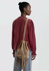 Frau mit dunkler Haut und Haaren zu einem Dutt gebunden, trägt ein rot langärmeliges Oberteil, hellblaue Jeans und eine beigefarbene Wildleder-Fransen-Schultertasche.