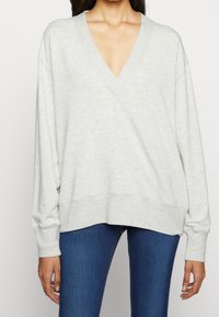 Sudadera oversized de color gris claro con escote en V profundo, mangas largas y puños de canalé; hecha de tejido suave, con un ajuste relajado.