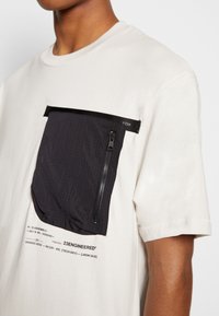 Bílá bavlněná košile s velkou černou kapsou na zip s mřížkovaným vzorem. Výrazné potiskované detaily pod kapsou. Krátké rukávy.