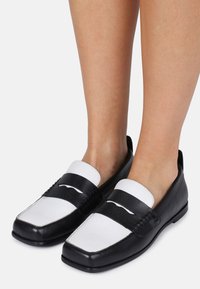Mocasines en blanco y negro con puntera cuadrada, un acabado de cuero suave y una correa en contraste a lo largo del empeine. Suelas planas, detalles mínimos.