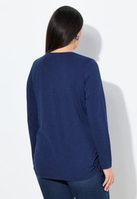 Haut à manches longues bleu marine avec col rond, texture douce et détails froncés sur les côtés, associé à un jean en denim sombre.