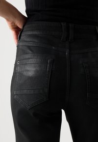 Svarta denimjeans har en åtsittande design med två bakfickor och synlig sömnad. Ytan har en svag lyster som framhäver strukturen.